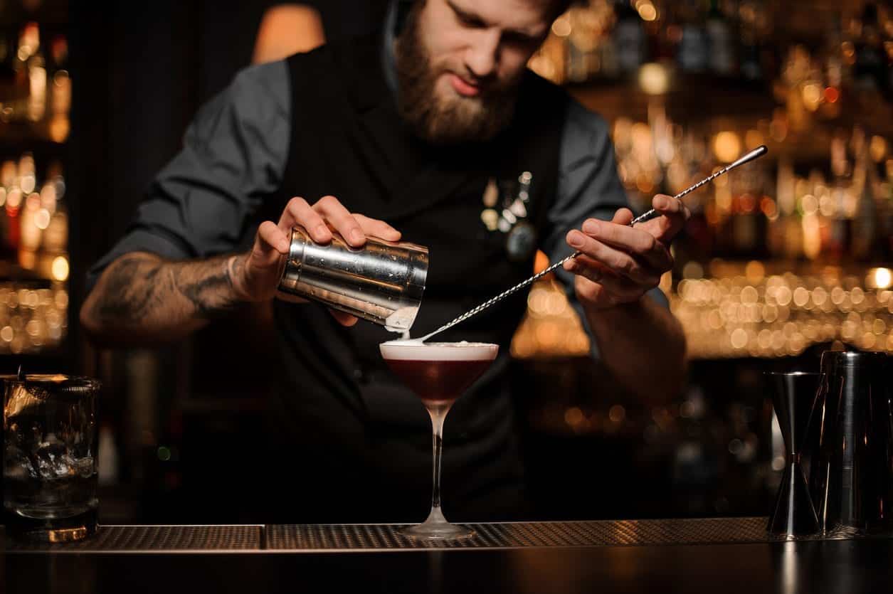 Brutal tattooed male bartender with beard pouring red alcohol cocktail using steel shaker and long bar spoon at bar counter