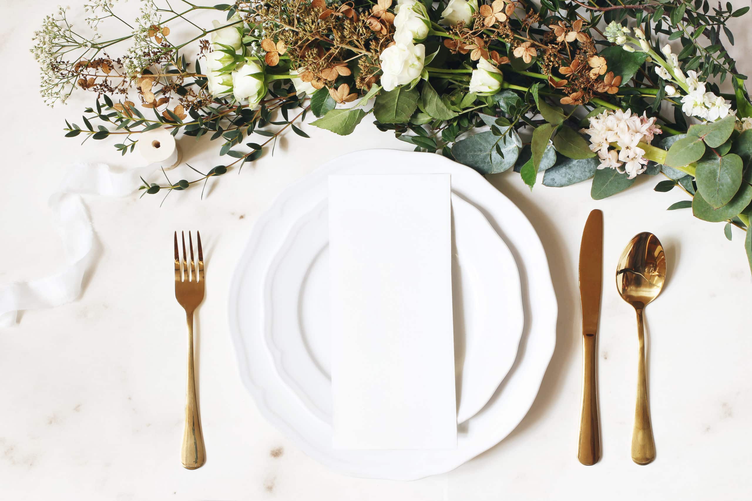 Festive wedding, birthday table setting with golden cutlery, silk ribbon, eucalyptus branches, hydrangea and white roses flowers. Blank card mockup. Restaurant menu concept, flat lay, top view