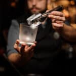 Professional bartender holding in hand an ice cube in tweezers putting it on the a cold matte cocktail glass on the bar counter on the blurred background.