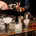 bartender carefully pours squeezed juice from jigger into steel shaker cup. Several glasses stand nearby.