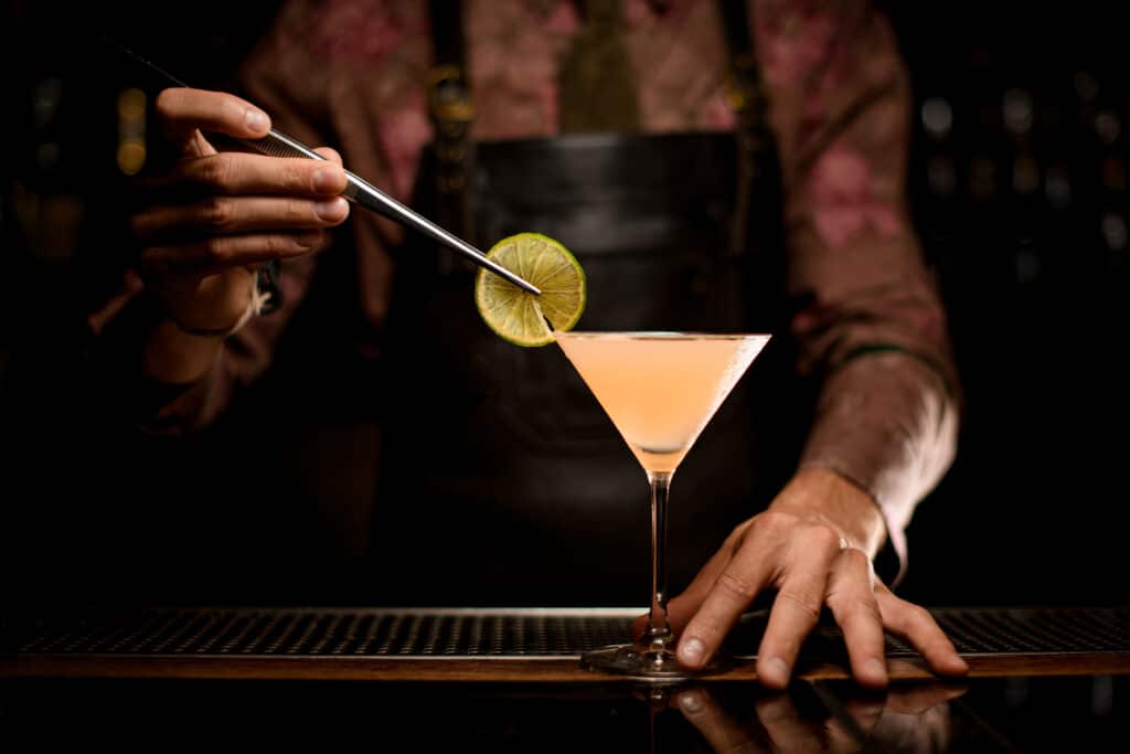 beautiful transparent martini glass with bright cocktail and hand of bartender holds slice of lemon with tweezers and decorates the glass with it