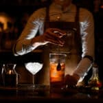 Male bartender in white shirt and leather apron stirring alcohol cocktail with a special bar spoon