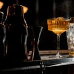 Glassy shaker and wineglass with ice brown alcoholic drink stand at bar counter. Several steel shakers stand nearby.