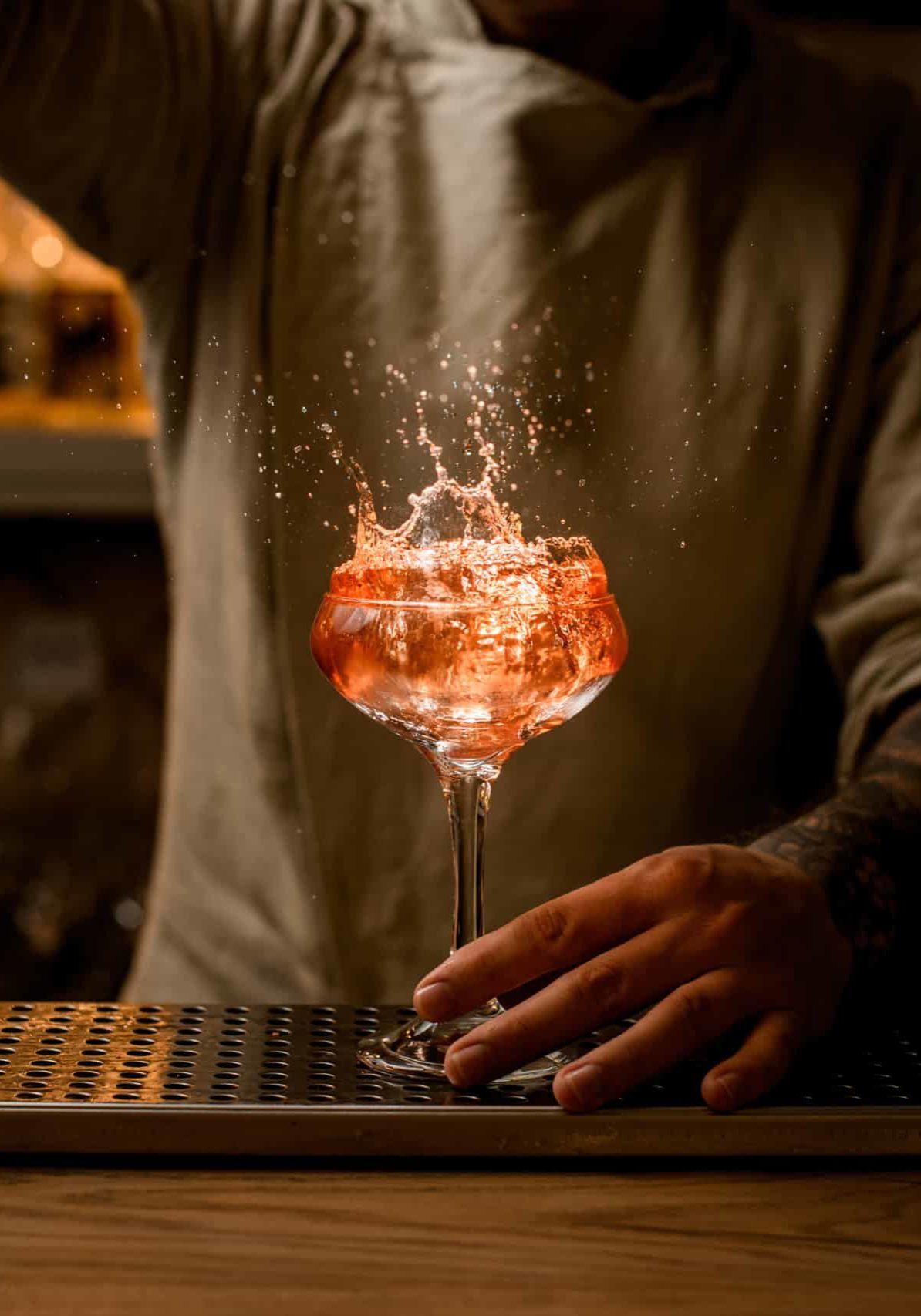 Barman's hand holds wine glass with splashes of bright cocktail which stands on the bar counter.