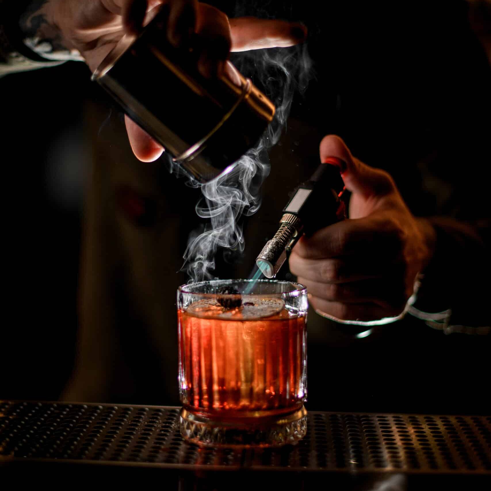 male bartender gently sprinkles white icing sugar on steaming glass with alcoholic cocktail on the bar counter