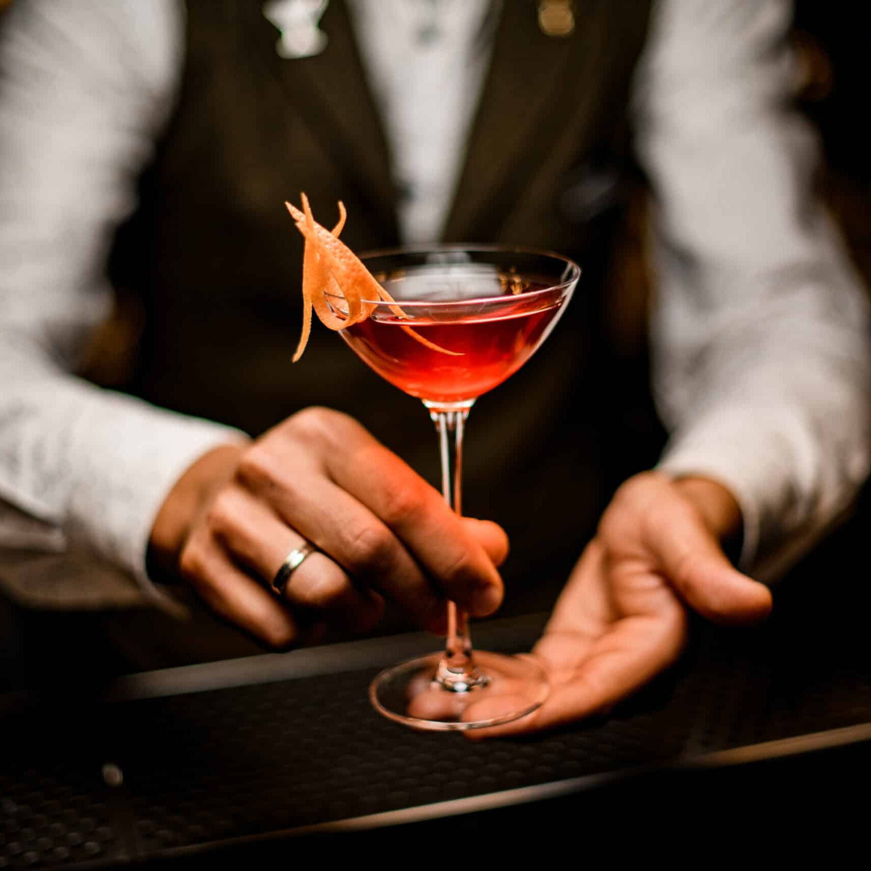 Close-up view of wine glass with trendy bright alcoholic cocktail in hands of male bartender. Blurred background