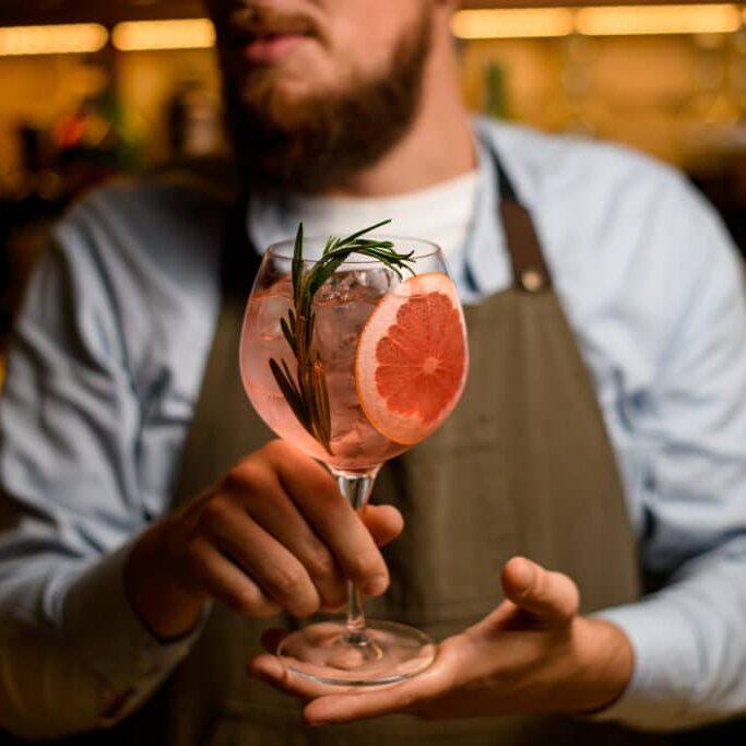 selective focus on goblet glass with cold drink decorating by a slice of grapefruit and fresh rosemary branch in male bartender hands