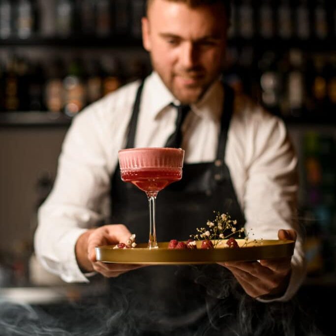 hand of barman holds round tray with raspberries and flowers and glass of pink foamy cocktail on it